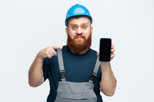 Серьезный молодой мужчина-строитель в защитном шлеме и униформе смотрит в камеру, показывая мобильный телефон на камеру, указывая на него на белом фоне