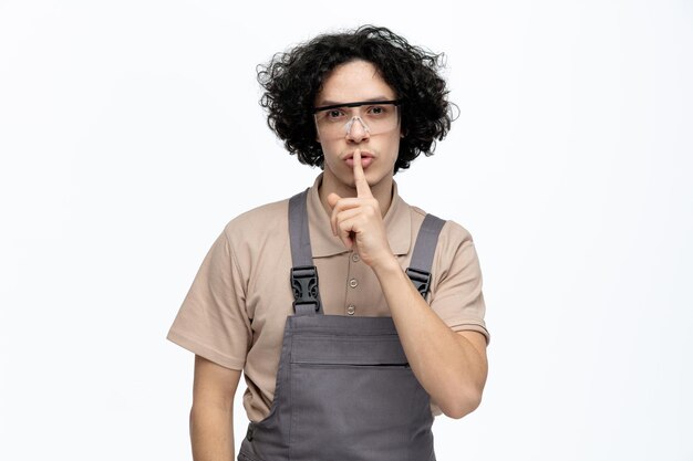 Serious young male construction worker wearing uniform and safety glasses looking at camera showing be silent gesture isolated on white background