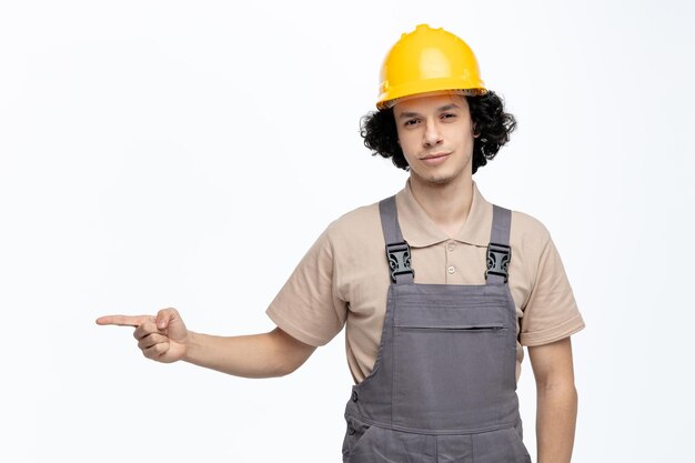 Serious young male construction worker wearing uniform and safety helmet looking at camera pointing to side isolated on white background