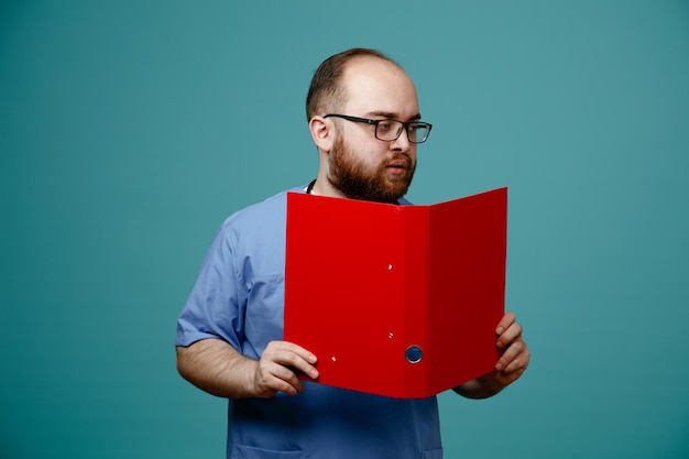 Serious young male nurse wearing glasses glasses nurse scrub and stethoscope around his neck holding folder looking at side isolated on blue background