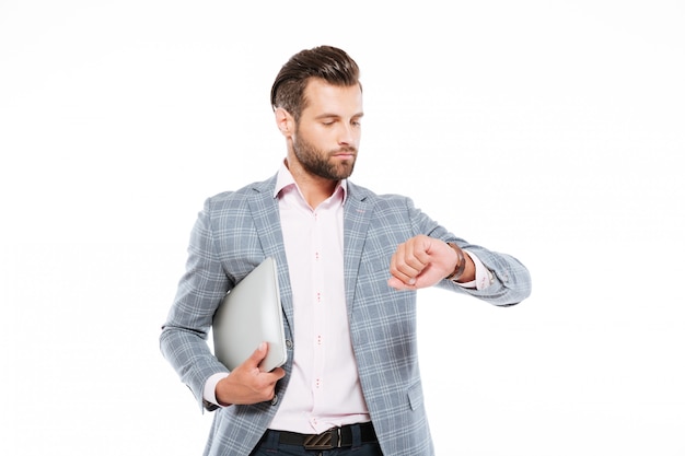 Serious young man holding laptop computer looking at watch.