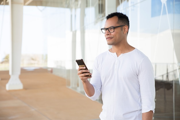 Serious young man holding phone in hands, looking aside