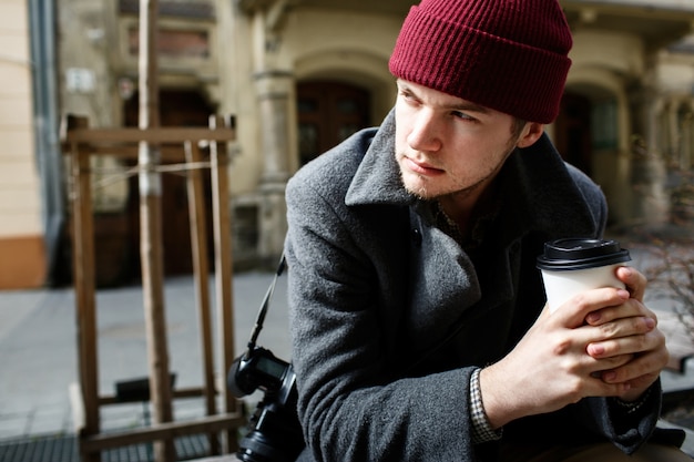 Serious young man holds paper cup of coffee sitting on the bench 