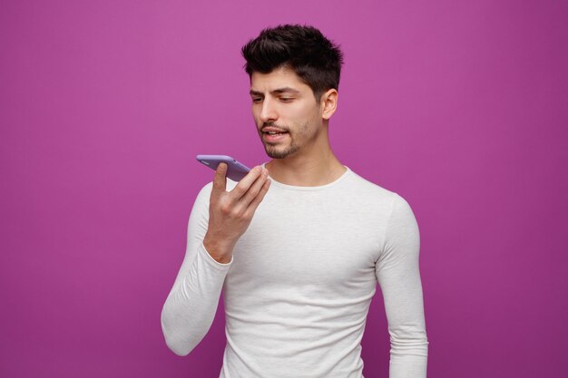 Serious young man looking down talking on phone using its microphone isolated on purple background