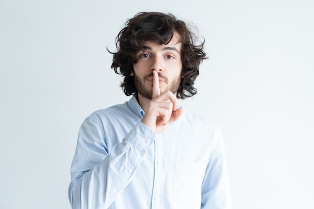 Serious young man making silence gesture and looking at camera