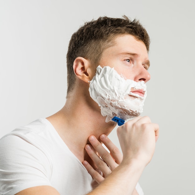 Serious young man shaving with blue razor against white background