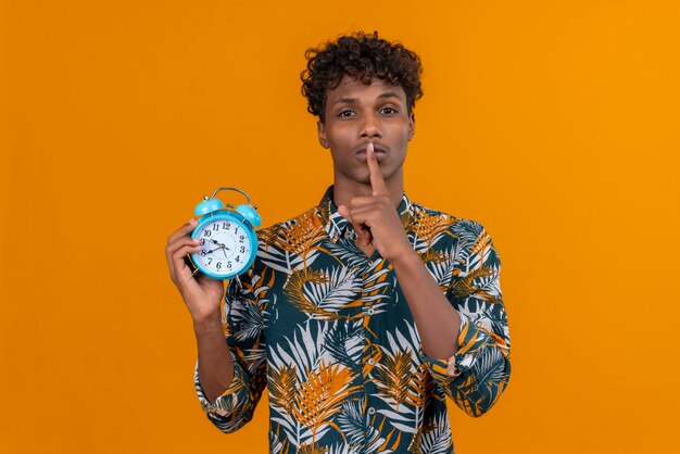 Serious young man in shirt showing shh gesture with index finger near mouth while holding alarm clock on an orange background