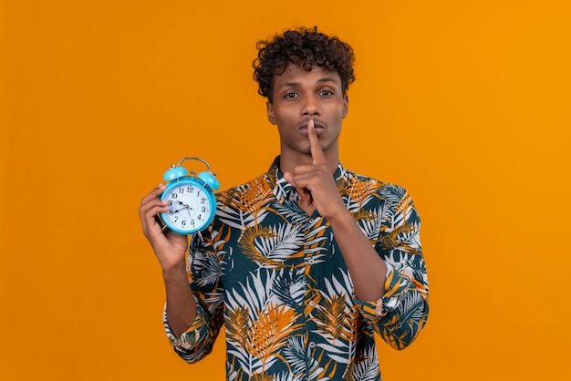 Serious young man in shirt showing shh gesture with index finger near mouth while holding alarm clock on an orange background