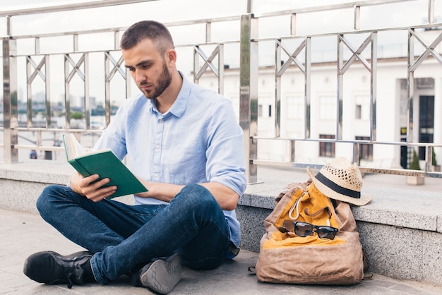 屋外で手すりの近くに座って本を読んで深刻な若い男