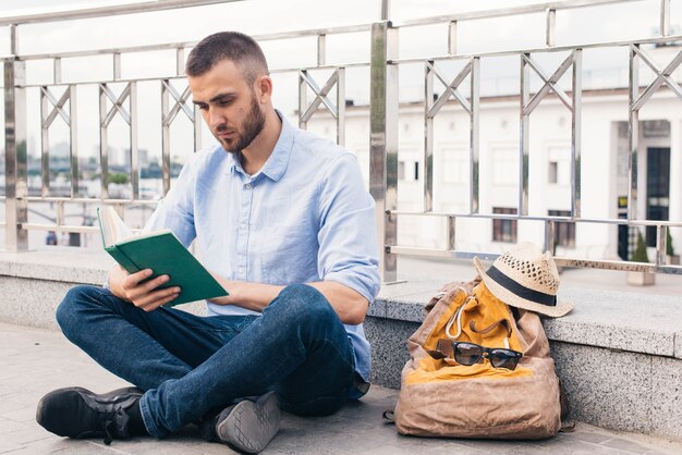 Serious young man sitting near railing at outdoors and reading book