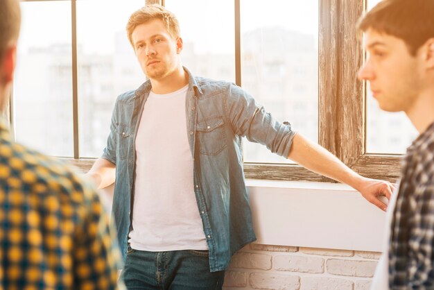 Serious young man standing near the window looking at his friend