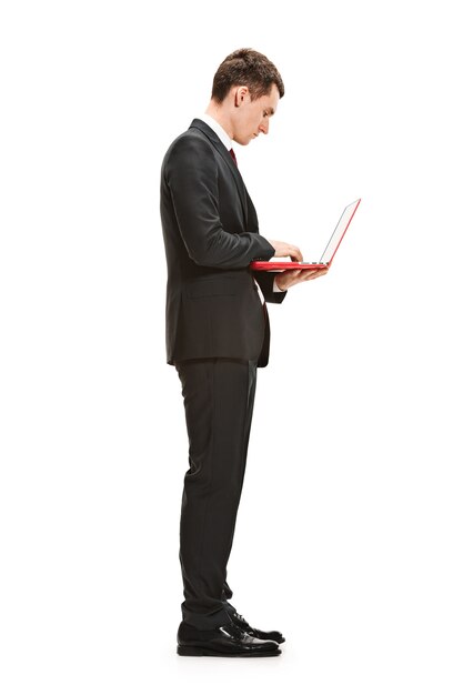 Serious young man in suit, red tie standing with laptop in office