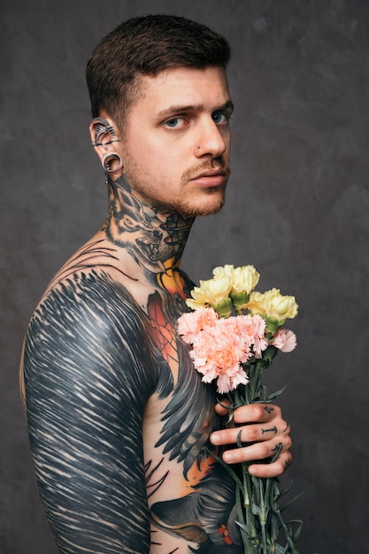 Free photo serious young man with pierced nose and ears holding carnation flowers in hand standing against gray backdrop