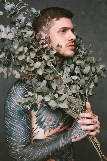 Serious young man with tattoo on his body holding branches of plant under his face