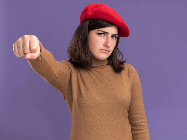 Serious young pretty caucasian girl with beret hat keeping fist isolated on purple wall with copy space