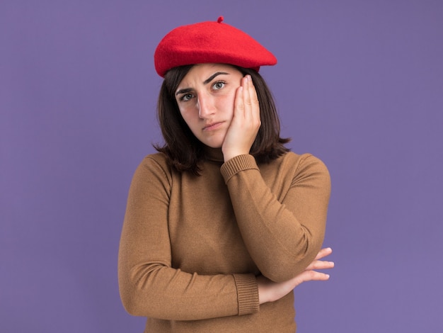 La giovane ragazza abbastanza caucasica seria con il cappello del berretto mette la mano sul fronte isolato sulla parete porpora con lo spazio della copia