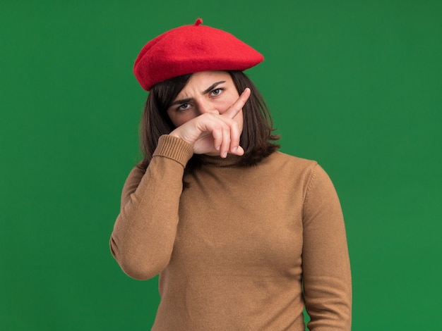ベレー帽の帽子をかぶった真面目な若いかなり白人の女の子は、コピースペースで緑の壁に分離された鼻に手を置きます