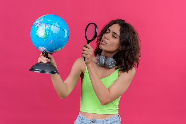 A serious young pretty woman with short hair in green crop top in headphones looking at globe steadily with magnifying glass