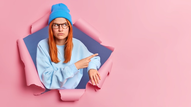 Foto gratuita grave giovane donna rossa che punta allo spazio della copia, indossa una felpa blu e un cappello. sfonda il muro di carta rosa.