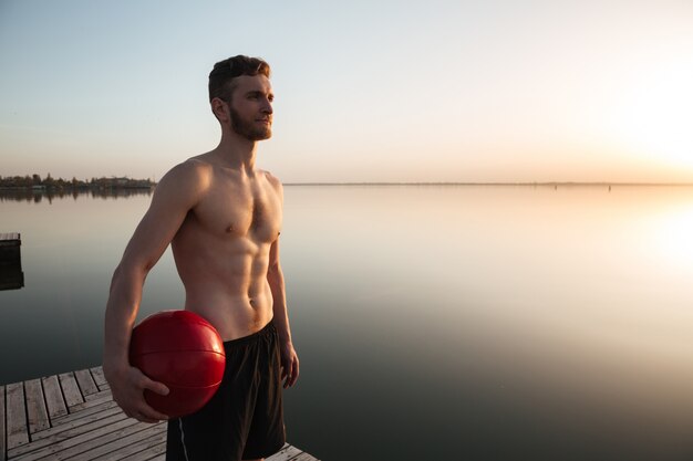 Serious young sportsman standing with ball at the beach.