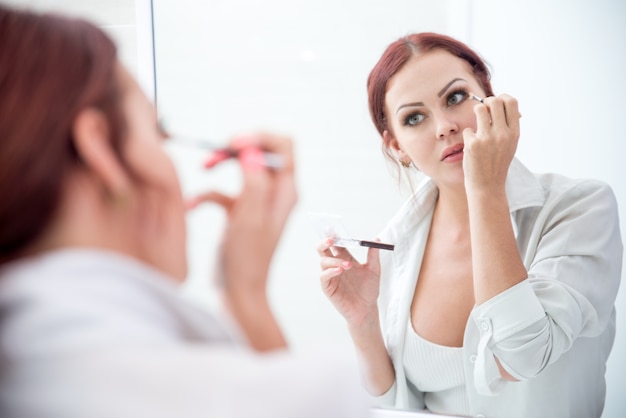 Serious young woman applying eyeshadow at mirror