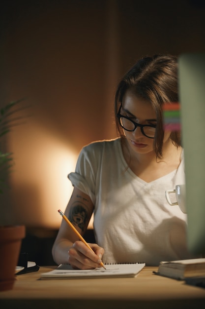Serious young woman designer sitting indoors at night writing notes