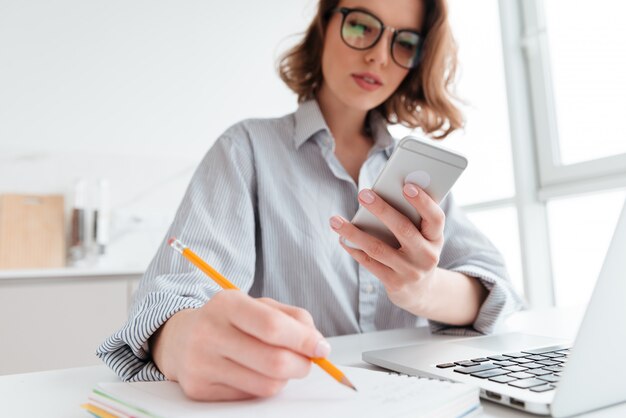 serious young woman in glasses holding mobile phone and taking notes while sitting in light apartment