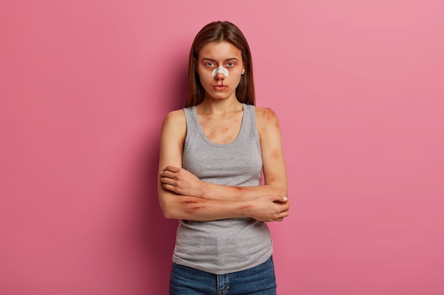 Serious young woman got nose bleed after insident, keeps arms folded over chest, being victim of domestic violence, beaten by someone, poses against pink wall, has bruised skin. Cruel sadism