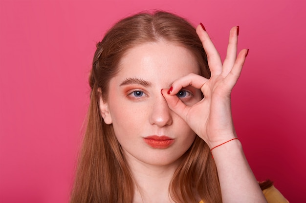 serious young woman, makes ok sign, cons her eye, expresses confidence, model poses on pink, being photographing. People and gesture concept.