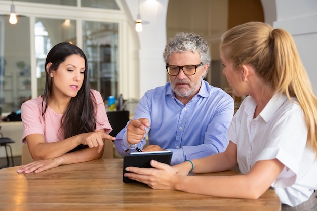 Serious young woman and mature man meeting with female professional, watching presentation on tablet, and pointing at screen