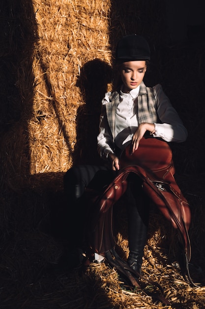 Free photo serious young woman sitting in barn with seddle