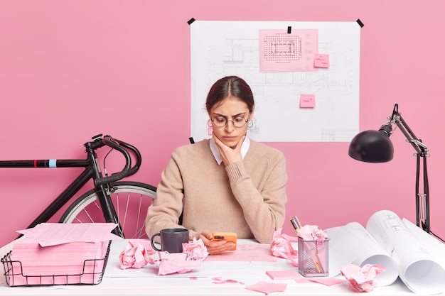 Serious young woman in spectacles makes sketches and blueprints in office using smartphone, poses at desktop against pink wall. Professional graphic designer develops new strategy