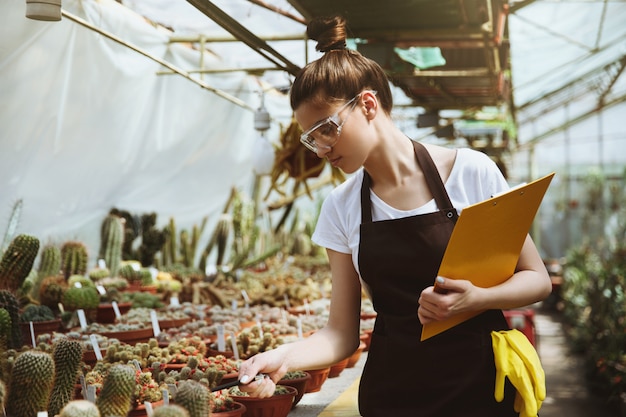 Serious young woman standing in greenhouse holding clipboard