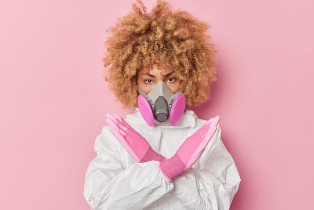 Serious young woman wears chemical protective suit and respirator makes stop gesture taboo sign doesnt allow to come in tries to prevent danger isolated over pink background Biohazard concept