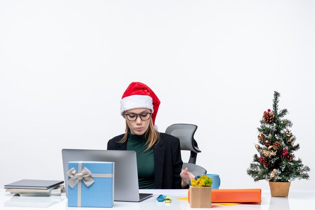 Serious young woman with santa claus hat and eyeglasses sitting at a table with a Xsmas tree and a gift on it in the office