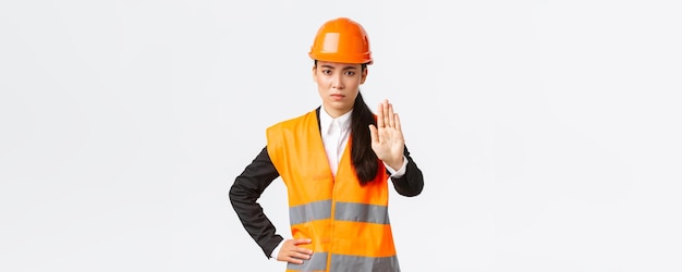 흰색 배경에 침입하는 행위를 금지하는 정지 제스처를 보여주는 안전 헬멧을 착용한 작업장에서 심각하게 실망한 아시아 여성 건축가 건설 관리자