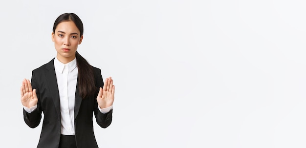 Серьезно выглядящая недовольная азиатская женщина-менеджер-предприниматель в черном костюме прекратит действие, запретит или ограничит что-то, поднимая руки в запрещающем жесте, говоря, что нет или достаточно белого фона