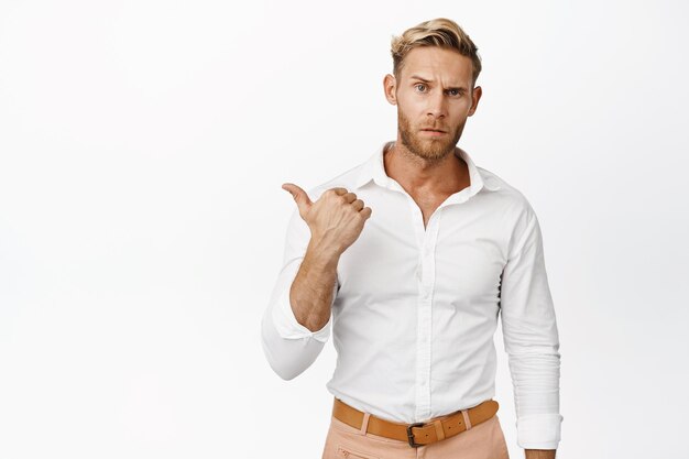 Seriouslooking macho man with blond hair and blue eyes frowning skeptical pointing finger left showing smth important white background