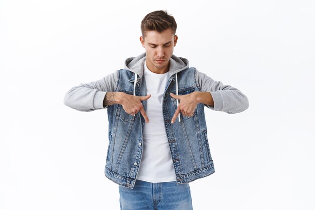 Seriouslooking young male student with blond hair denim jacket looking and pointing fingers down with concerned judgemental expression see something important at bottom promo