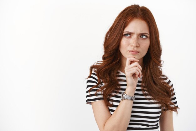 Seriouslooking young redhead woman scheming something look suspicious and hesitant having doubts express trong disbelief as pondering information touch chin frowning look sideways thoughtful