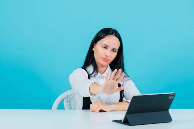 Seriously blogger girl is showing stop gesture by sitting in front of tablet camera on blue background