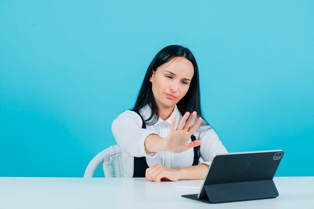 真剣にブロガーの女の子は青い背景の上のタブレットカメラの前に座って停止ジェスチャーを示しています