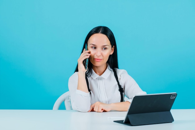 Seriously blogger girl is talking on phone by sitting in front of tablet camera on blue background