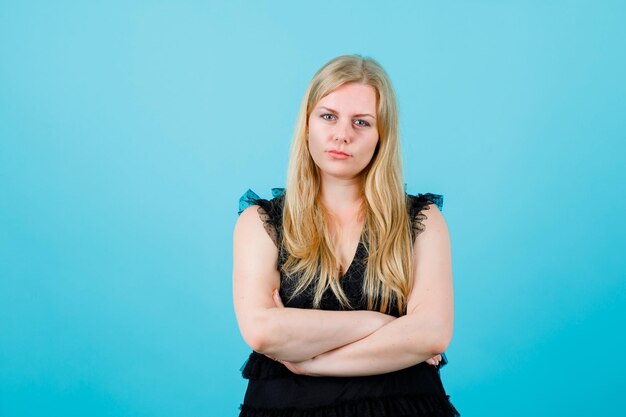 Seriously blonde girl is looking at camera by crossing arms on blue background
