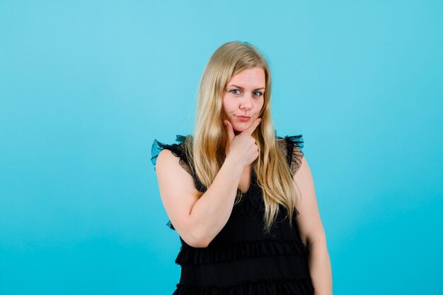 Seriously blonde girl is looking at camera by holding hand on chin on blue background