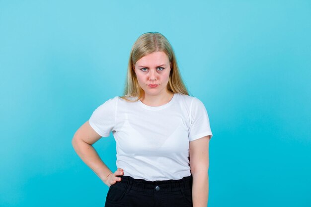 Seriously blonde girl is looking at camera by putting hand on waist on blue background