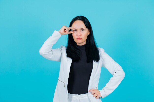 Seriously girl is holding fingers around eye and putting other hand on waist on blue background