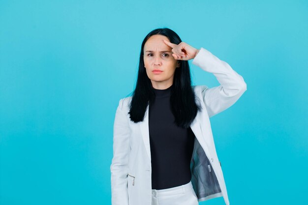 Seriously girl is looking at camera by holding forefinger on forehead on blue background