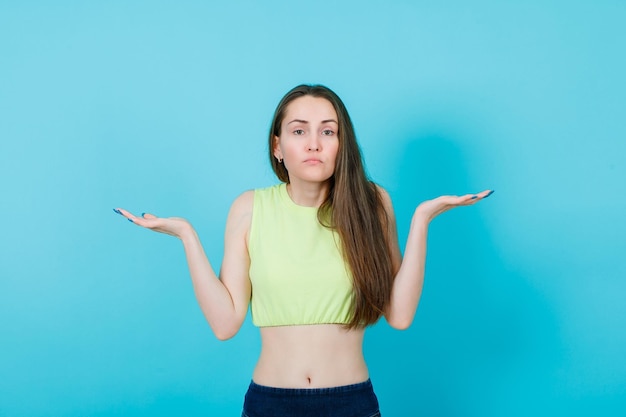 Seriously girl is looking at camera by opening wide her arms on blue background