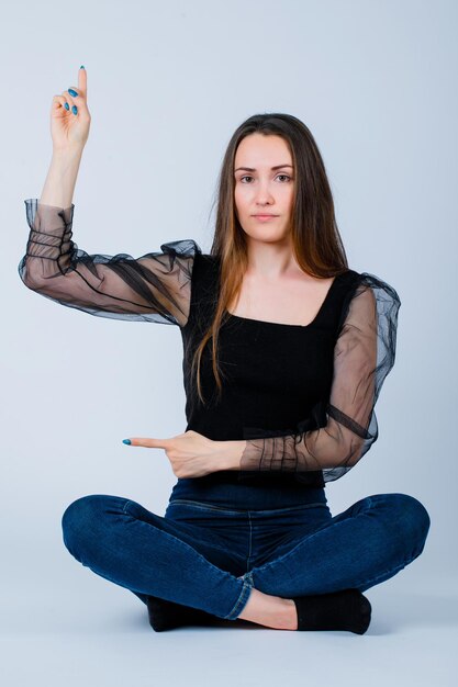 Seriously girl is pointing up and left with forefingers by sitting on floor on white background